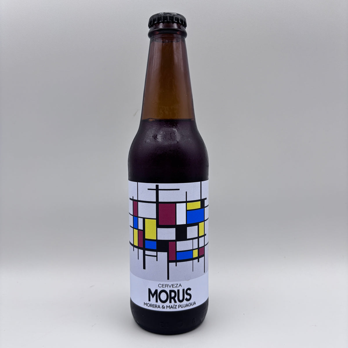 Botella de Cerveza Morus elaborada con morera y maíz pujagua, con etiqueta blanca decorada con un diseño geométrico de cuadrícula en colores sobre un fondo neutro.