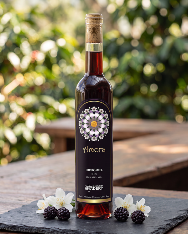Botella de hidromiel marca Amora de Costa Rica Meadery sobre una mesa de madera al aire libre, decorada con zarzamoras frescas y flores blancas en la base.